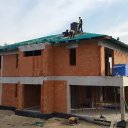 Dwukondygnacyjny dom w trakcie budowy: murowane ściany z czerwonej cegły, częściowo wykonana konstrukcja dachu z zieloną membraną, robotnicy na dachu montują komin.