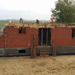 Budowa domu jednorodzinnego: murowane ściany z pustaków ceramicznych, drewniane szalunki stropu, widoczni pracownicy w odzieży roboczej. Tło: pola uprawne.