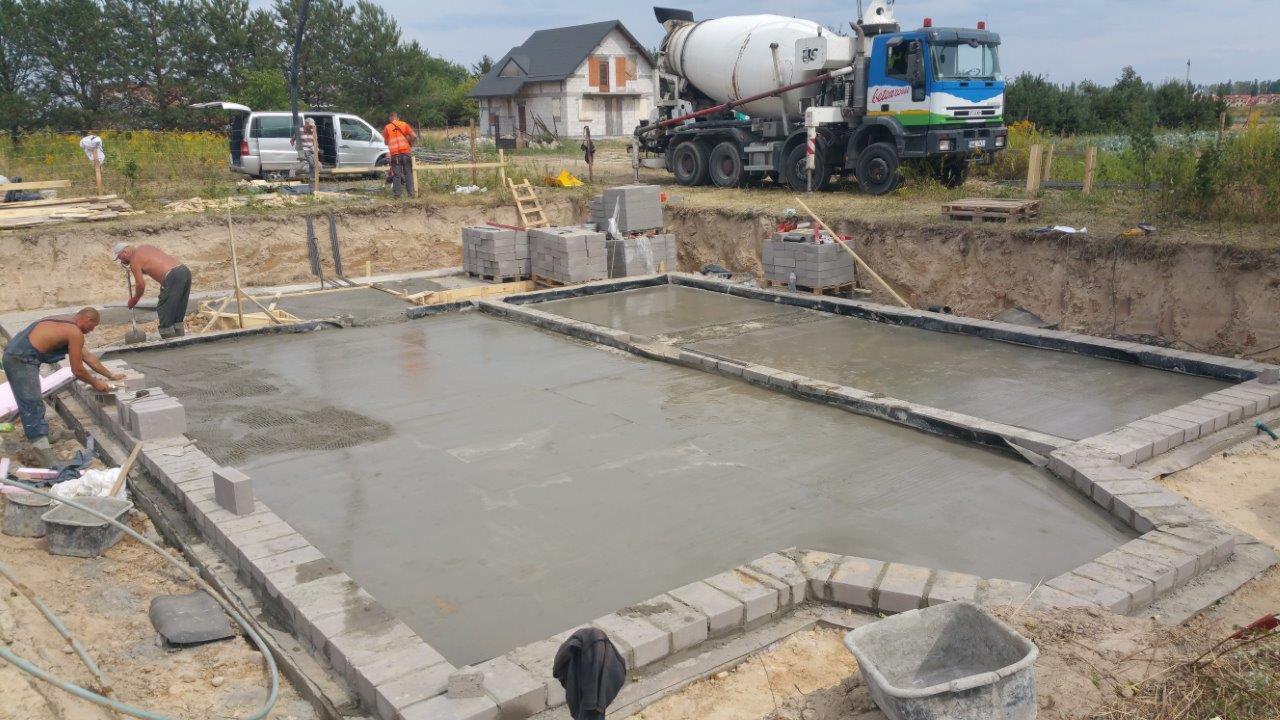 Wylewanie fundamentów pod budynek jednorodzinny. Widoczny betonowy fundament w szalunku z bloczków betonowych. W tle betoniarka i ekipa budowlana.