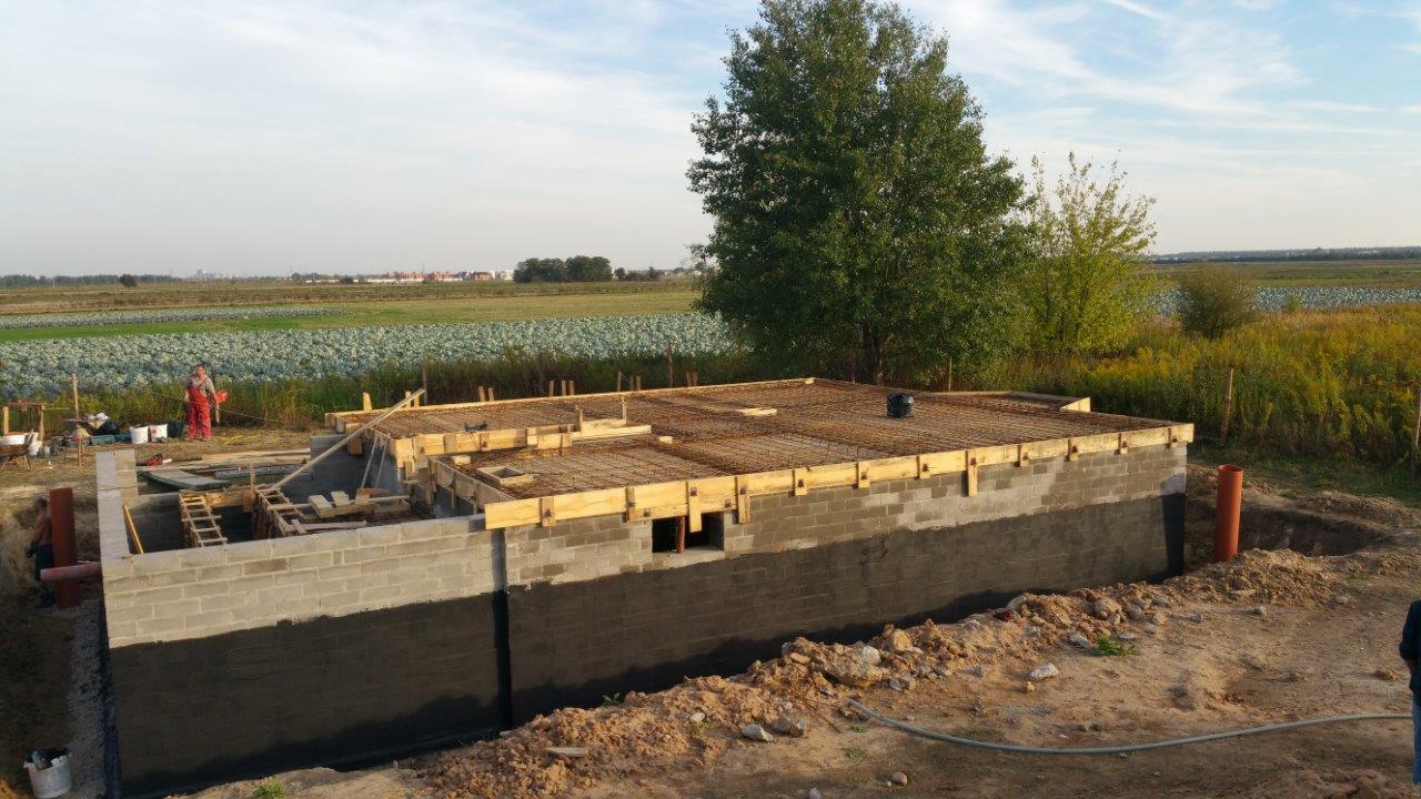 Budowa fundamentów z betonowych bloków i zbrojenia, widok z zewnątrz. W tle pole i drzewa. Prace budowlane w toku, widoczny pracownik w czerwonym kombinezonie.