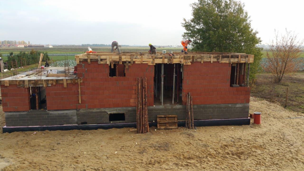 Budowa domu jednorodzinnego: murowane ściany z pustaków ceramicznych, drewniane szalunki stropu, widoczni pracownicy w odzieży roboczej. Tło: pola uprawne.