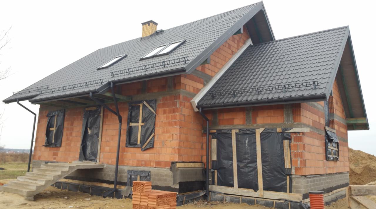 Niedokończony dom z czerwonej cegły z szarym dachem, czarnymi rynnami i oknami zabezpieczonymi folią. Widoczne fundamenty i schody wejściowe.