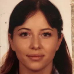 Portret młodej kobiety z ciemnymi włosami i brązowymi oczami, uśmiechająca się delikatnie, na jasnym tle. Widoczne delikatne cieniowanie i naturalny makijaż.