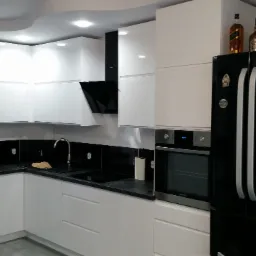 Biała, nowoczesna kuchnia z czarnym blatem i lodówką. Górne szafki zaokrąglone, na lodówce butelki alkoholu i model żaglowca. Oświetlenie sufitowe.