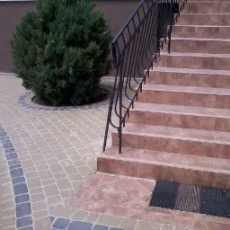 Kaskadowe schody z terakoty prowadzą w górę, obok ścieżki z kostki brukowej w odcieniach beżu i szarości. Uroku dodaje krzew iglasty.