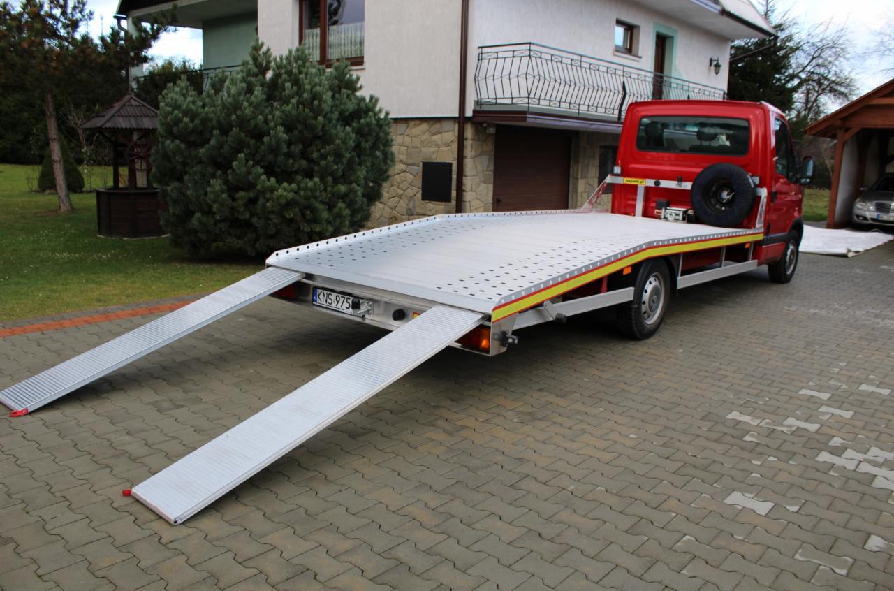 Czerwona laweta z rozłożonymi rampami na brukowanym podjeździe, na tle domu z jasną elewacją i ogrodem. Widoczny numer rejestracyjny i koło zapasowe.