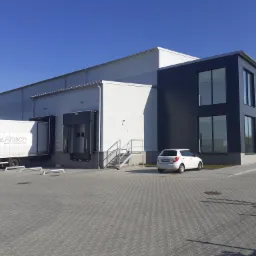 Nowoczesna hala magazynowa z rampami rozładunkowymi, białą elewacją z płyt warstwowych i ciemnoszarą częścią biurową z dużymi oknami, na parkingu stoi biały samochód osobowy, a do rampy podjeżdża...