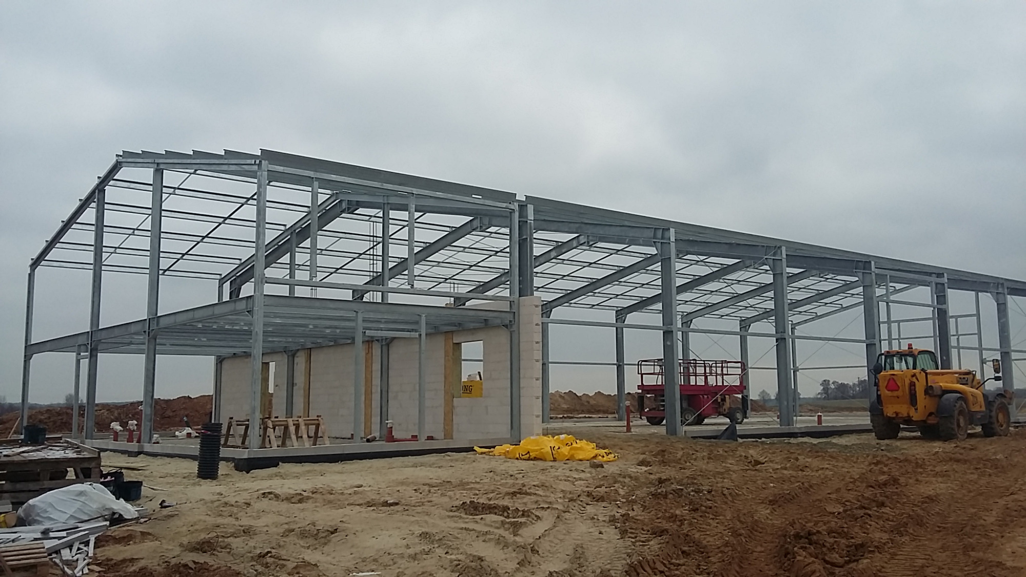 Stalowa konstrukcja hali produkcyjno-magazynowej w trakcie budowy, widoczny szkielet budynku, ściany z bloczków, żółte worki z materiałami budowlanymi oraz żółta ładowarka teleskopowa na placu budowy.