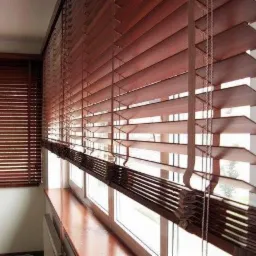 Drewniane żaluzje poziome w odcieniach brązu, częściowo zasłonięte, zamontowane w oknach. Widoczne sznurki regulacyjne i lamele. W tle kolejne okno z żaluzjami.