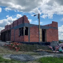 Wznoszenie ścian z czerwonej cegły na budowie domu jednorodzinnego w Myślenicach, widoczny samochód osobowy i malowniczy krajobraz w tle pod błękitnym niebem.