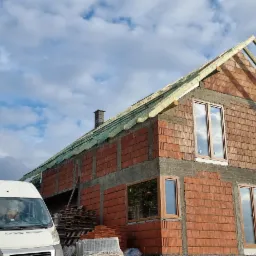 Wznoszony dwukondygnacyjny budynek z czerwonej cegły ceramicznej z zamontowanymi oknami, widoczne elementy konstrukcji dachu, na pierwszym planie biały samochód dostawczy, w tle pagórkowaty krajobraz.