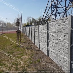 P.P.H.U. WAWMAR MAREK WAWRZONOWSKI - Długi, szary mur o fakturze kamienia, biegnący wzdłuż boiska sportowego, z widoczną konstrukcją wysokiego napięcia w tle.