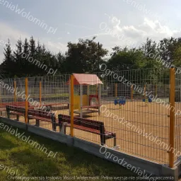 Plac zabaw otoczony siatką z metalowych paneli na betonowej podmur&oacute;wce, widoczne ławki i urządzenia do zabawy.