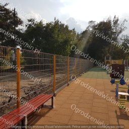 P.P.H.U. WAWMAR MAREK WAWRZONOWSKI - Ogrodzenie panelowe z pomarańczowymi słupkami na placu zabaw, widoczna ławka i urządzenia dla dzieci w tle, w tle drzewa i niebo.
