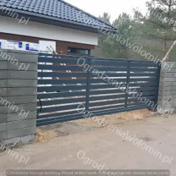 Nowoczesna posesja z przesuwną, metalową bramą wjazdową z poziomymi panelami, osadzoną na betonowych słupkach z bloczków, widok z zewnątrz.