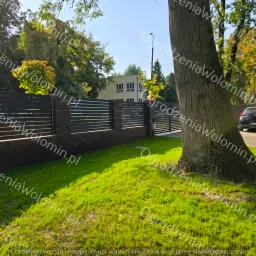 Nowoczesne ogrodzenie z poziomych, ciemnych paneli metalowych i ceglanych słupków, widok zza pnia drzewa na tle budynku i zielonego trawnika.