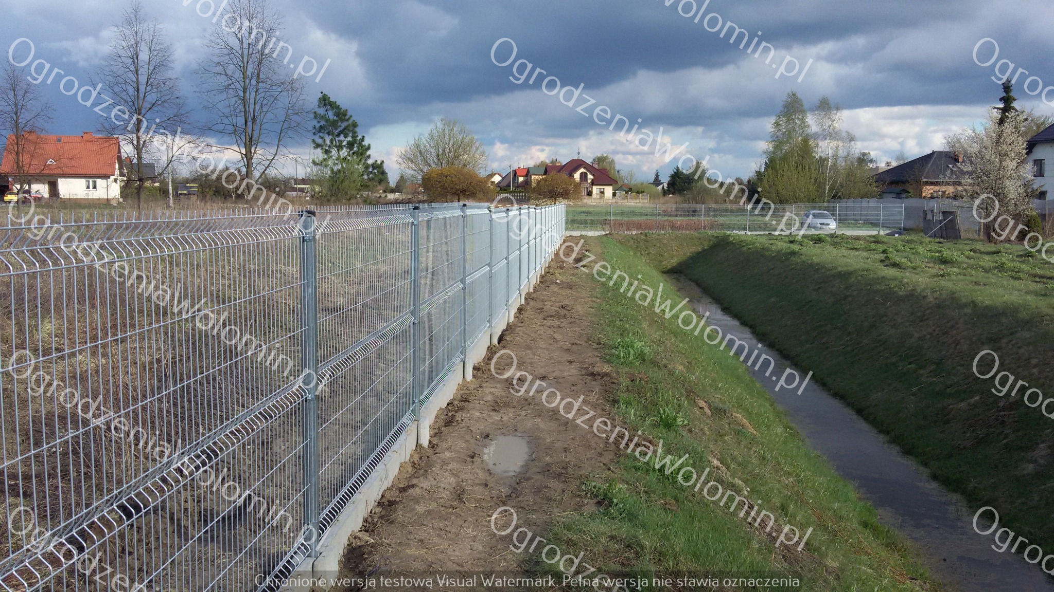 Długi, szary panel ogrodzeniowy z betonową podmurówką, oddzielający posesję od rowu melioracyjnego, widok z perspektywy ziemi w pochmurny dzień.