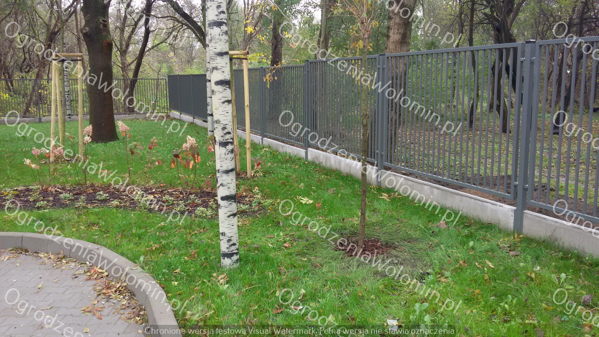 Szare metalowe ogrodzenie z pionowych prętów, osadzone na betonowej podmurówce, widoczne na tle parku z drzewami i zielonym trawnikiem, z młodymi drzewkami podpartymi drewnianymi stelażami.