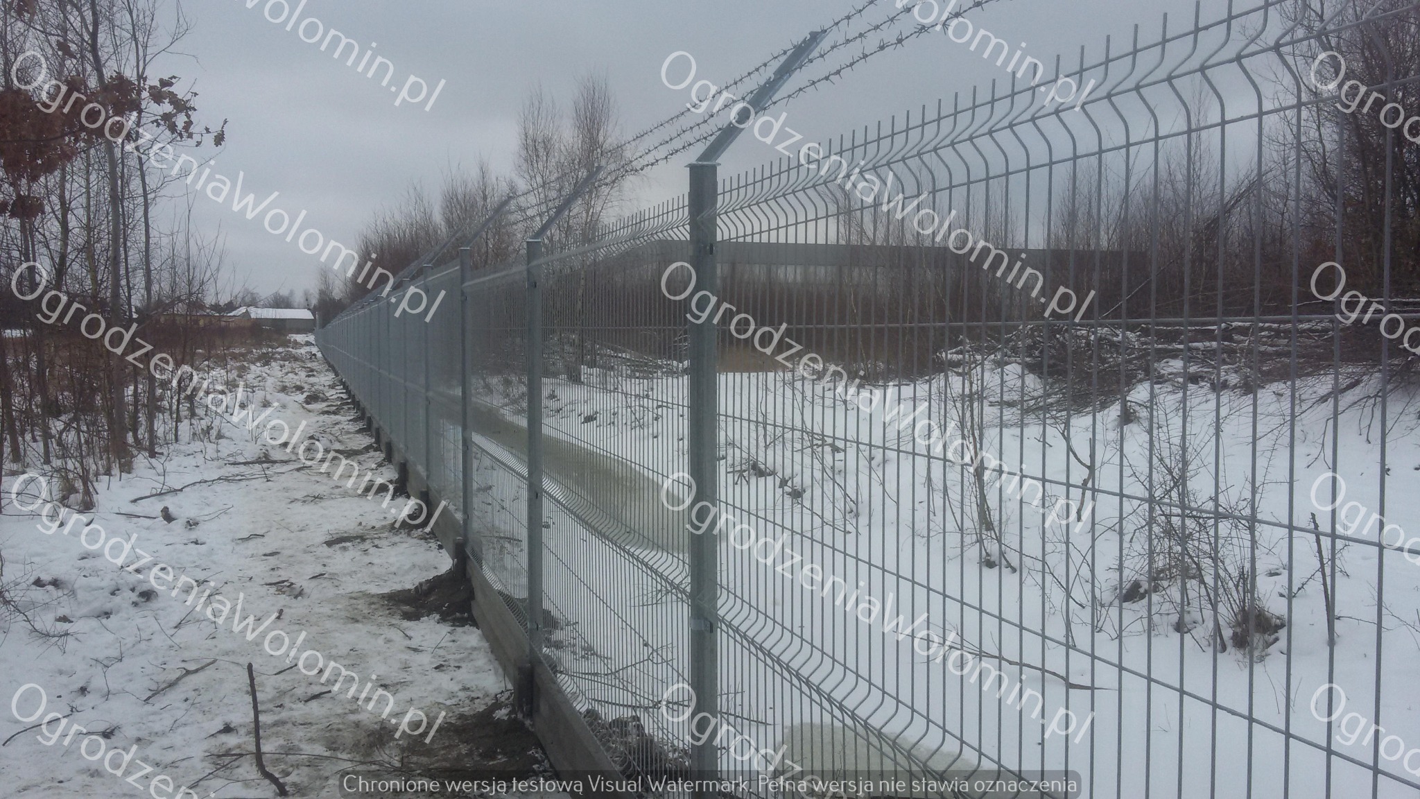 Długi, szary system zabezpieczeń z drutu kolczastego nad siatką, rozciągnięty wzdłuż zasypanego śniegiem terenu z drzewami w tle, widok z perspektywy.