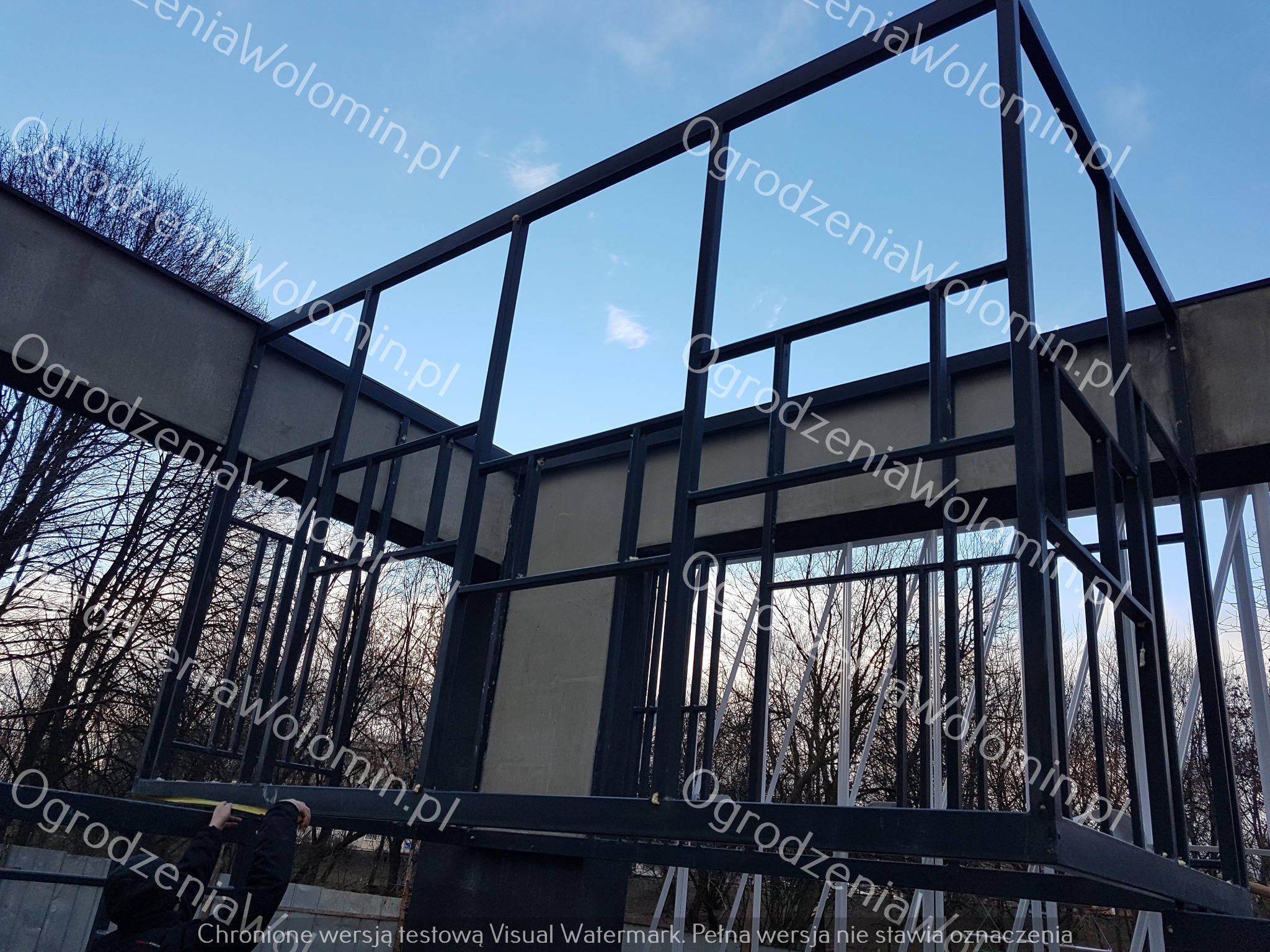 Konstrukcja metalowa ogrodzenia w trakcie montażu, widoczny pracownik z miarką, na tle drzew i błękitnego nieba.