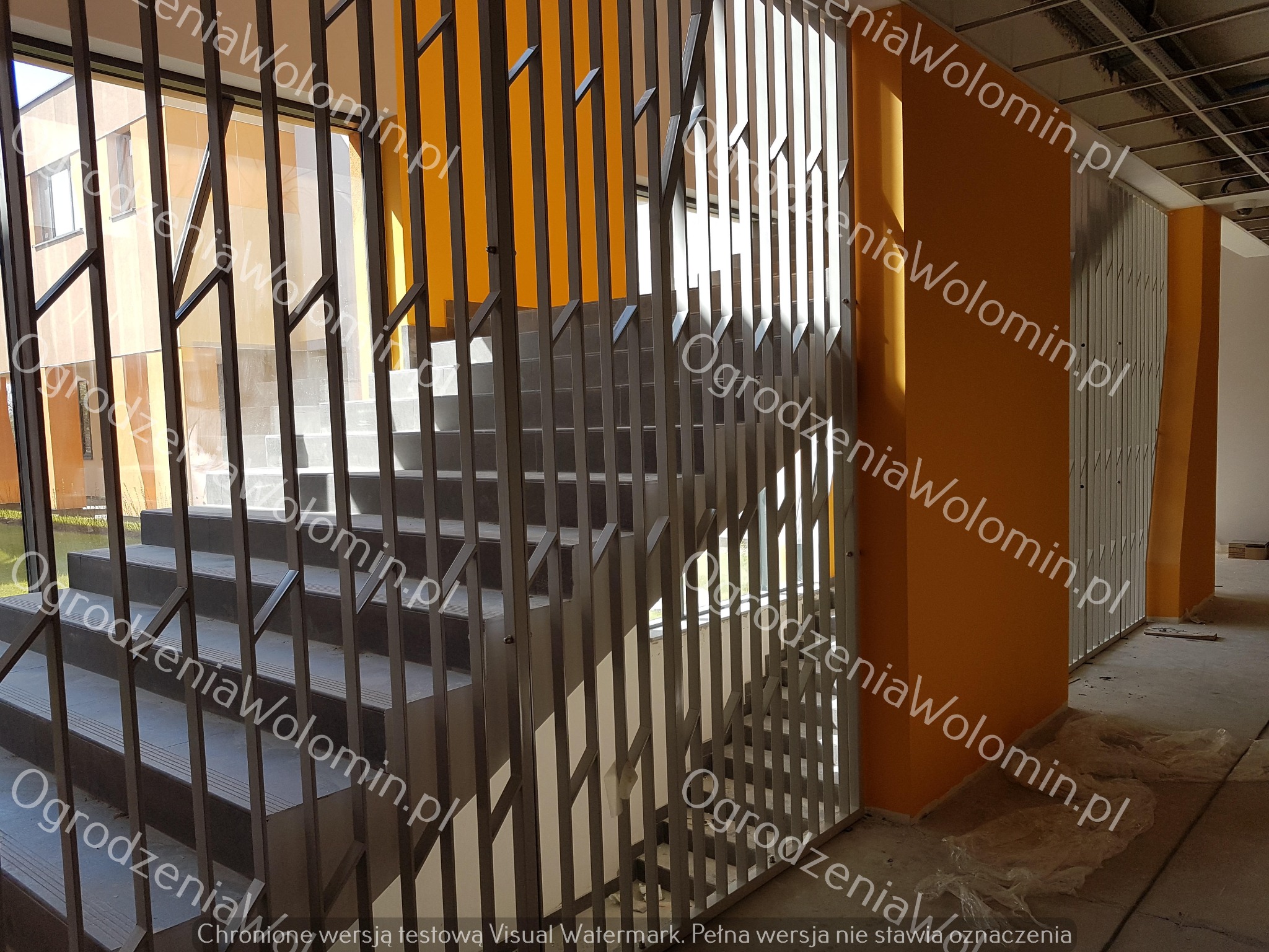 Metalowa balustrada schodowa o nowoczesnym designie w budynku w Wołominie, z widocznymi schodami i pomarańczową ścianą w tle.