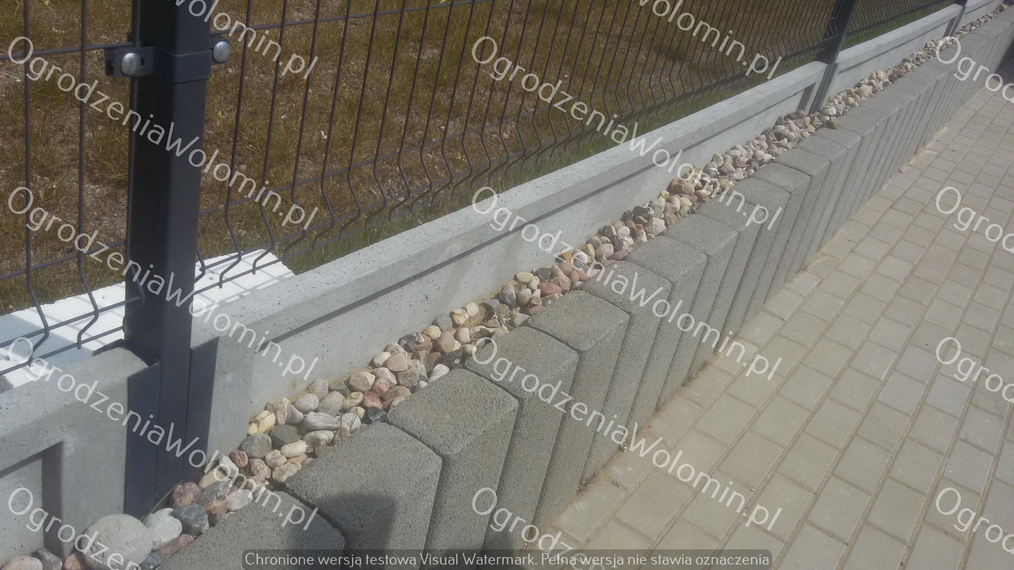 Fragment ogrodzenia z siatki panelowej na betonowej podmurówce, ozdobionej kamieniami rzecznymi, obok ułożona kostka brukowa.