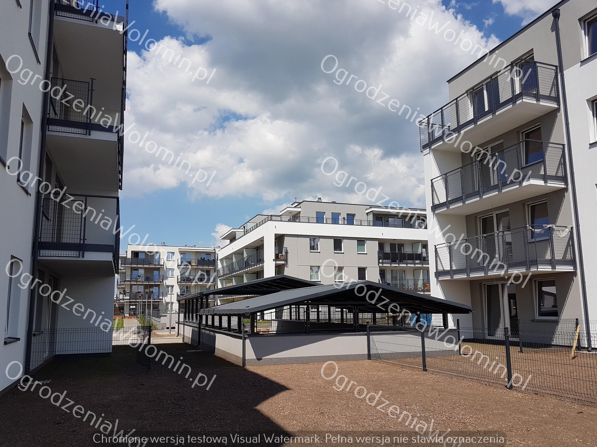 Nowoczesne osiedle z widoczną siatką ogradzającą teren, w tle zadaszona konstrukcja, błękitne niebo z chmurami.