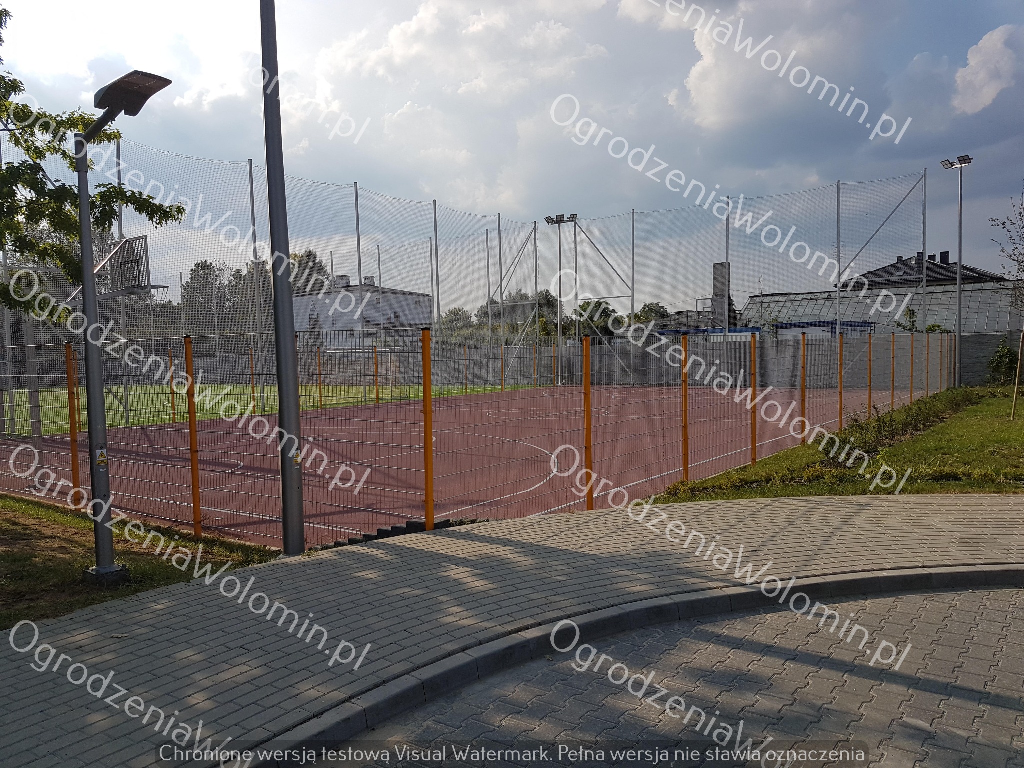 Ogrodzenie boiska sportowego z siatki i paneli, widok z chodnika z kostki brukowej, oświetlenie boiska.