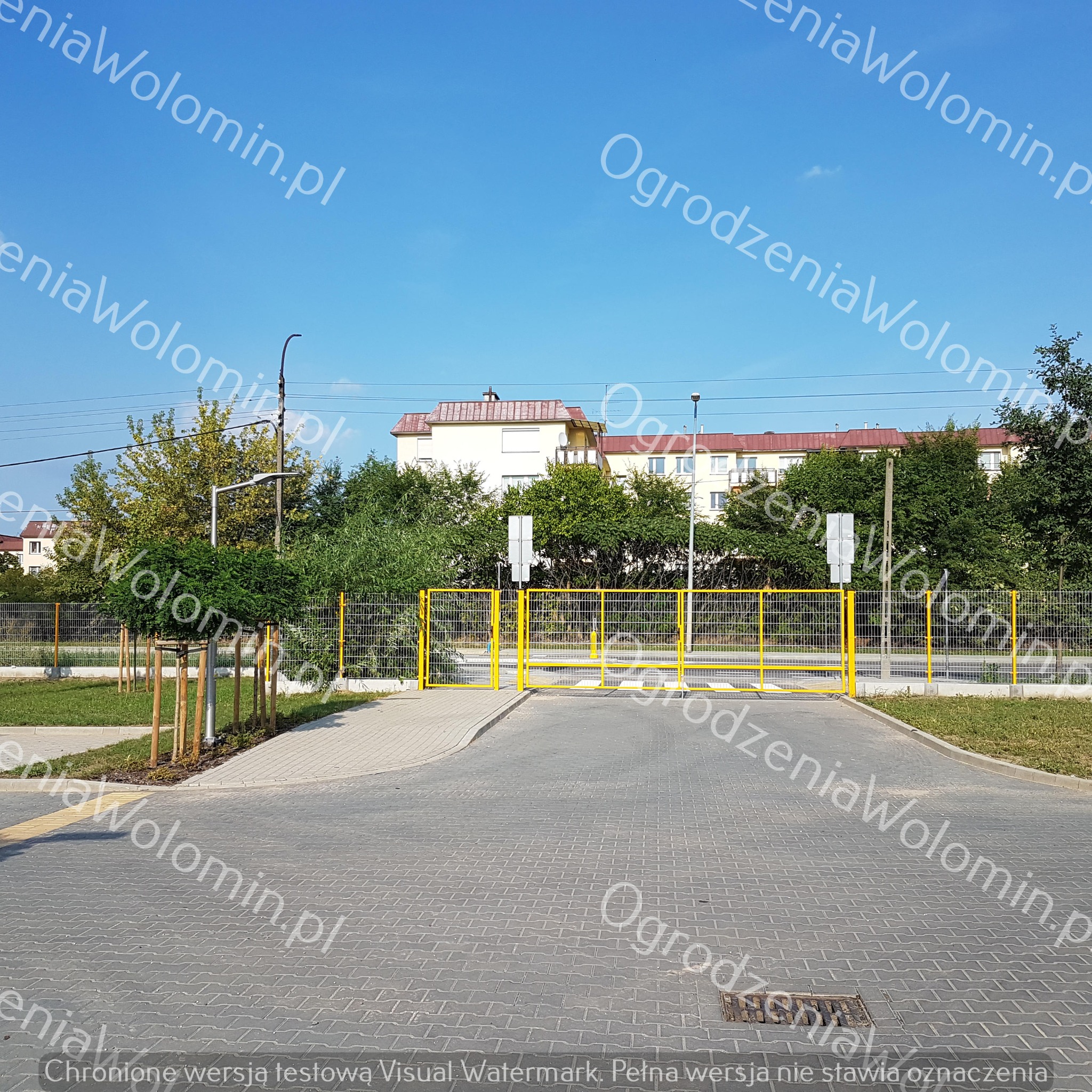 Żółta brama wjazdowa z siatki na osiedlu mieszkaniowym, widok z zewnątrz na kostkę brukową i budynki w tle.