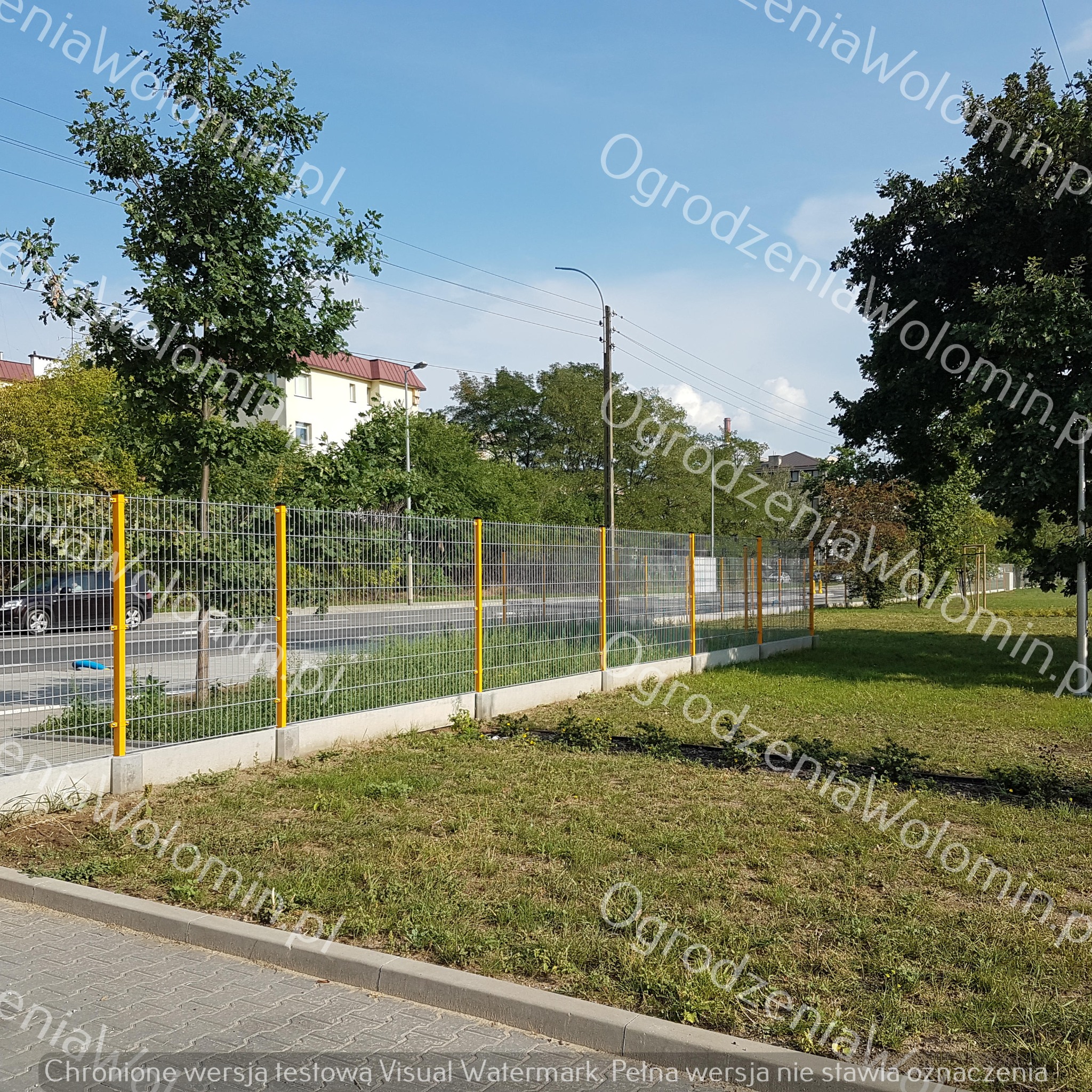 Długi odcinek ogrodzenia z siatki z żółtymi słupkami, osadzonego na betonowej podmurówce, oddzielający chodnik od trawnika i zabudowań mieszkalnych w tle, w słoneczny dzień.