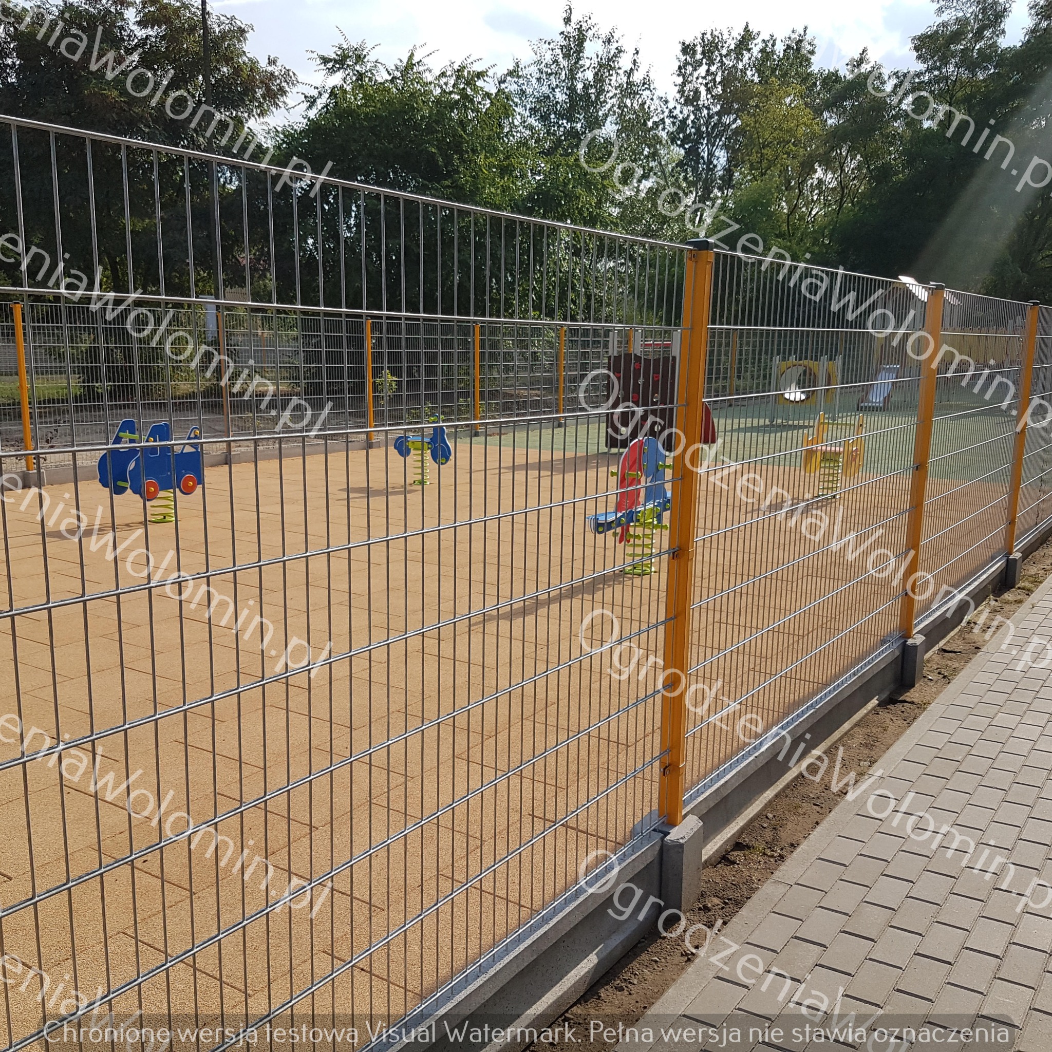 Metalowe ogrodzenie panelowe z żółtymi słupkami otaczające plac zabaw z kolorowymi zabawkami na piaskowej nawierzchni, widok z zewnątrz na kostkę brukową.