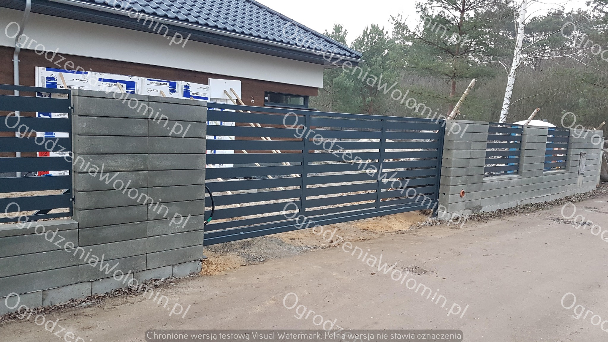 Nowoczesna posesja z przesuwną, metalową bramą wjazdową z poziomymi panelami, osadzoną na betonowych słupkach z bloczków, widok z zewnątrz.