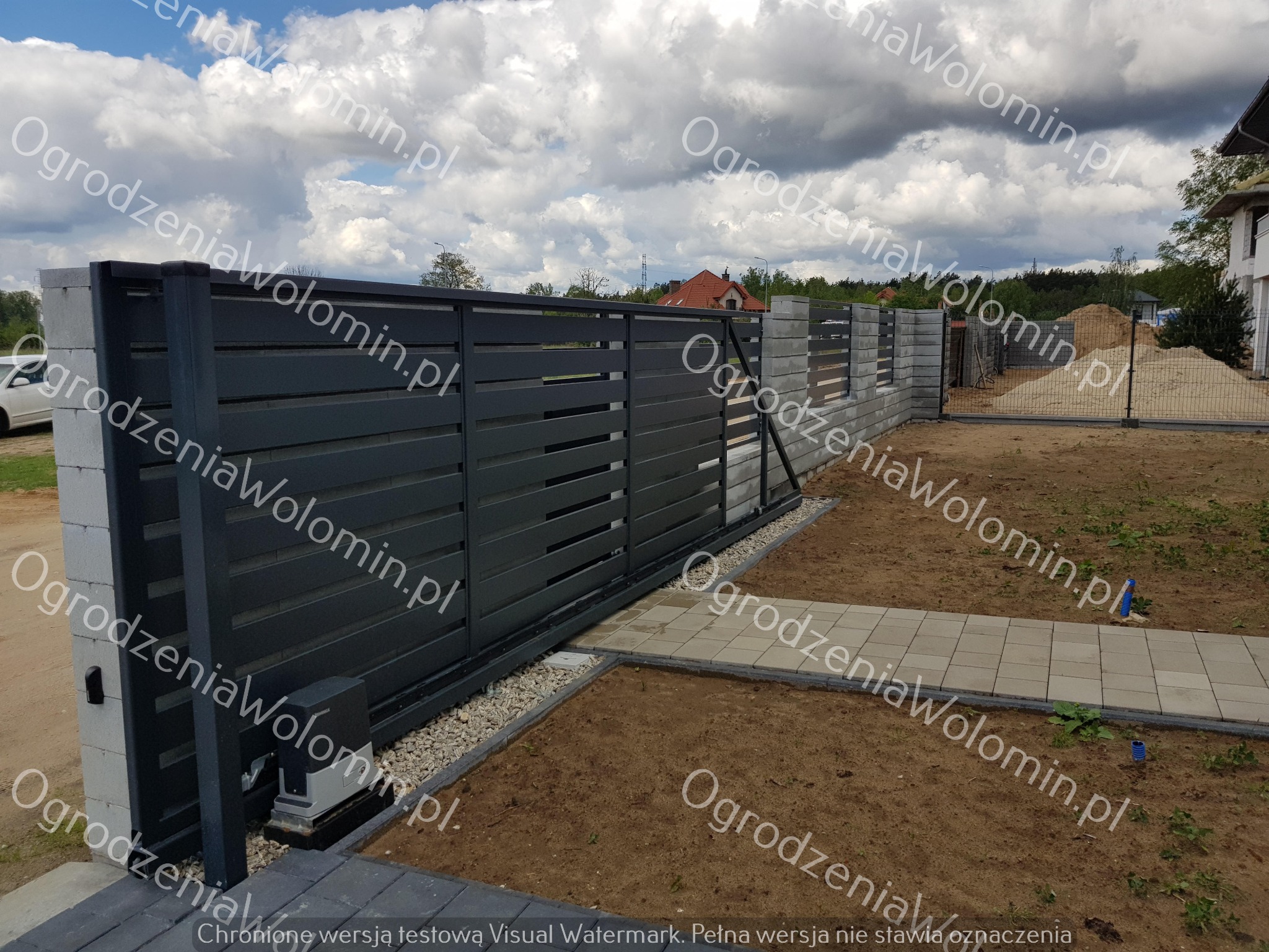 Nowoczesne, poziome przęsła metalowe w kolorze grafitowym, osadzone na betonowych słupkach. Widoczny automat do bramy przesuwnej i ścieżka z kostki brukowej. W tle siatka ogrodzeniowa i kopiec piasku.