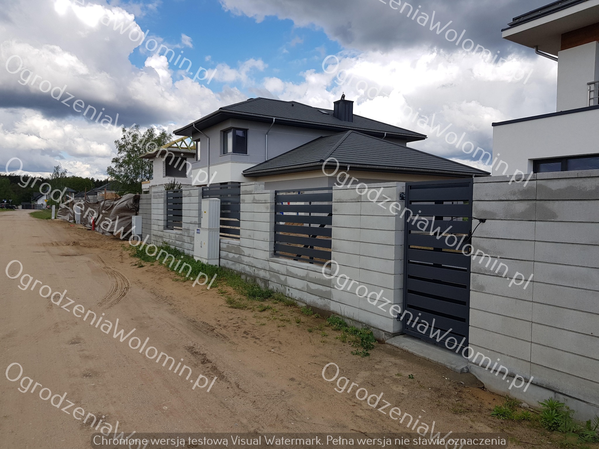 Nowoczesne ogrodzenie z szarego betonu architektonicznego z poziomymi, metalowymi przęsłami i bramą wjazdową, na tle domu z ciemną dachówką i pochmurnego nieba.