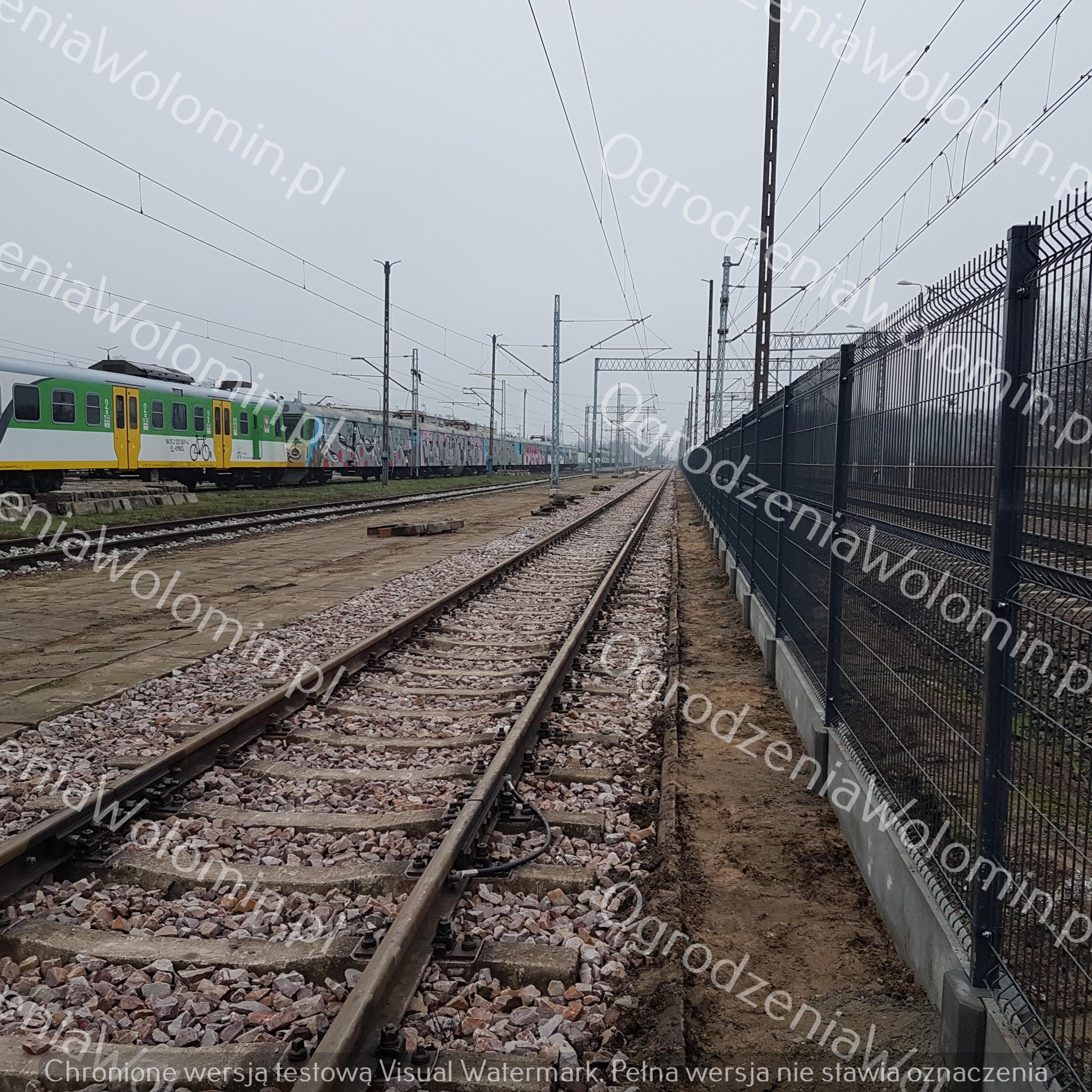 Nowoczesne, czarne ogrodzenie panelowe z podmurówką betonową, oddzielające tory kolejowe od terenu przyległego, z widocznym pociągiem w tle.
