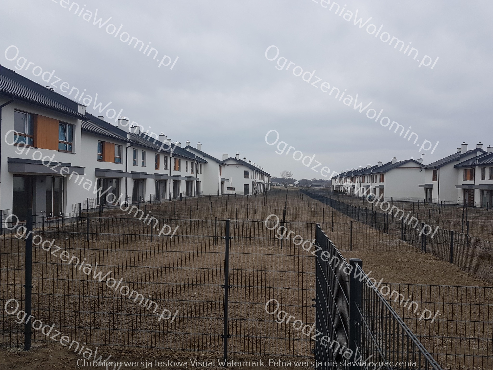 Montaż metalowego ogrodzenia panelowego na osiedlu domów szeregowych, widok na przygotowany teren z wbitymi słupkami i częściowo zamontowanymi przęsłami.