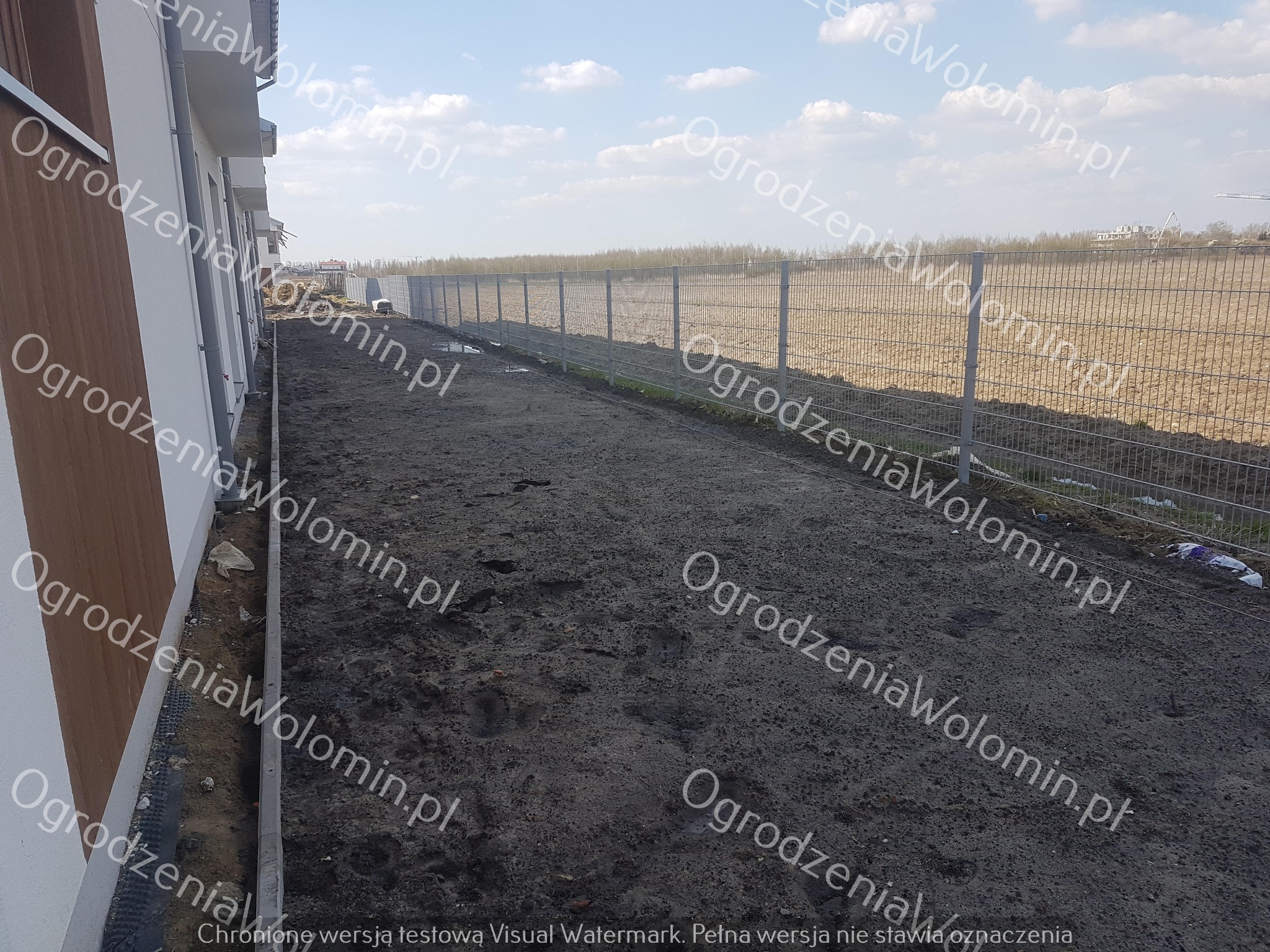 Długie, szare ogrodzenie z siatki panelowej, widoczne z perspektywy gruntu, oddzielające teren budowy od pola. W tle widoczne budynki i drzewa.