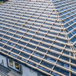 Wysoki kąt na konstrukcję dachu z łat drewnianych i folii dachowej Ventia NEO z widocznymi oznaczeniami, na tle domów i zieleni.