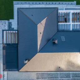 Widok z góry na nowoczesny dom z ciemnoszarą dachówką ceramiczną, przylegający taras z pergolą, zadbany trawnik i wybrukowane ścieżki.