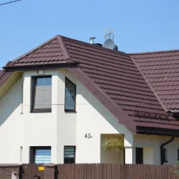 Dom z brązowym dachem z dachówki ceramicznej i białymi ścianami, widoczne okna PCV i antena satelitarna na dachu. Ujęcie z poziomu ulicy, słoneczny dzień.