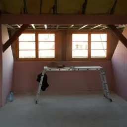 Poddasze w trakcie adaptacji: ściany obłożone różową płytą, widoczne drewniane belki stropowe, okna z drewnianą ramą, rozstawiona drabina aluminiowa i butelka wody mineralnej w rogu pomieszczenia.