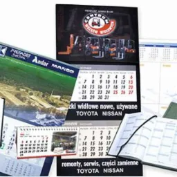 Różnorodne kalendarze firmowe: biurkowe, ścienne, książkowe, z logo firm i grafikami, prezentujące usługi i produkty. Wyeksponowane daty i nazwy marek.