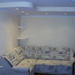 Jasny salon z narożnikiem i podwieszanym sufitem z oświetleniem punktowym. Na ścianie półki. Prosty wystrój, neutralne kolory, ciepłe światło.