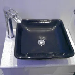 Błyszcząca, czarna, kwadratowa umywalka z chromowaną armaturą na blacie z białego lastryko. Białe płytki w tle i dozownik mydła po prawej.