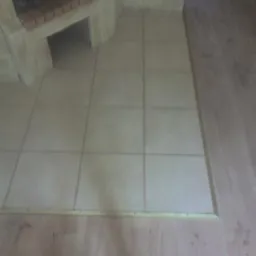 Beżowe płytki podłogowe ułożone przy kominku, przechodzące w drewnianą podłogę oddzieloną listwą. Widoczny fragment kominka z ceglanym wykończeniem.