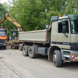 Dwa samochody ciężarowe Mercedes i koparka podczas załadunku ziemi na poboczu drogi w słoneczny dzień. Widoczne drzewa w tle.