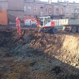 Pomarańczowo-biała koparka Atlas i wywrotka na placu budowy, wykop pod fundamenty. Budynek w tle. Tablice reklamowe widoczne po lewej stronie.