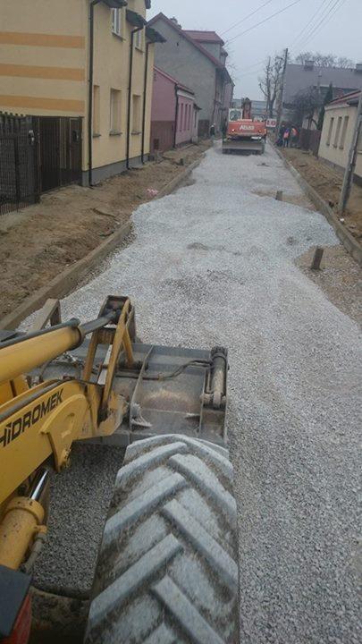 Żółta ładowarka 'Hidromek' na tle drogi z tłucznia w zabudowie miejskiej. W tle czerwona maszyna budowlana i budynki.