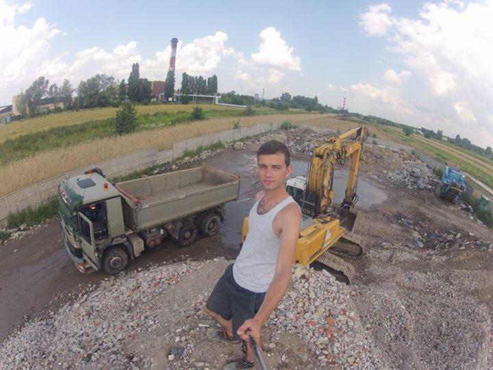 Mężczyzna robi selfie na tle wywrotki i koparki na placu budowy z gruzem, w tle komin i zieleń, szeroki kąt.