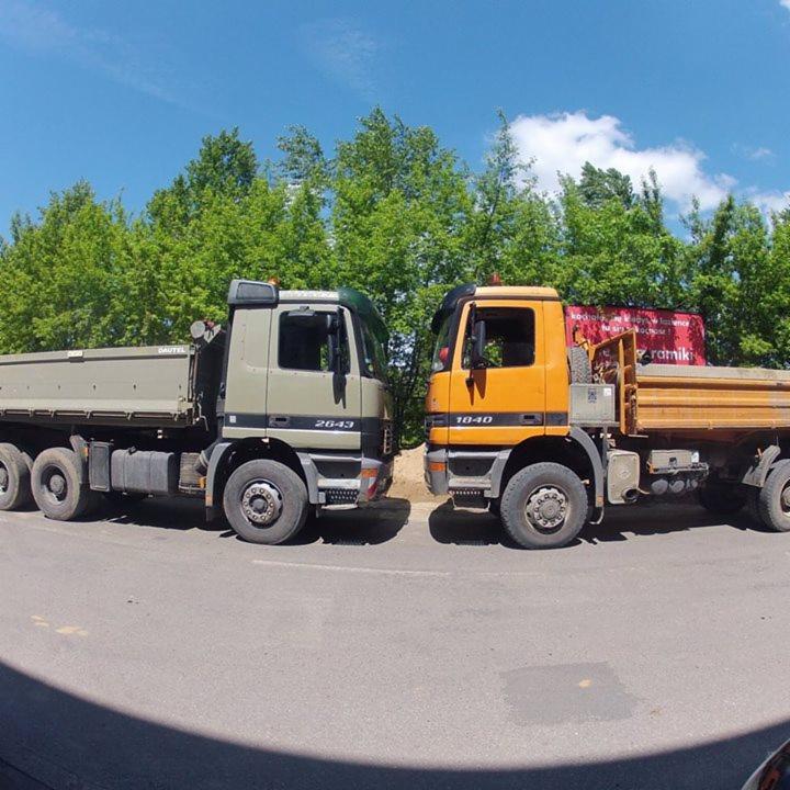 Dwa samochody ciężarowe (wywrotki) różnych marek zaparkowane na asfalcie, z zielonymi drzewami w tle i błękitnym niebem. Widok z niskiej perspektywy.